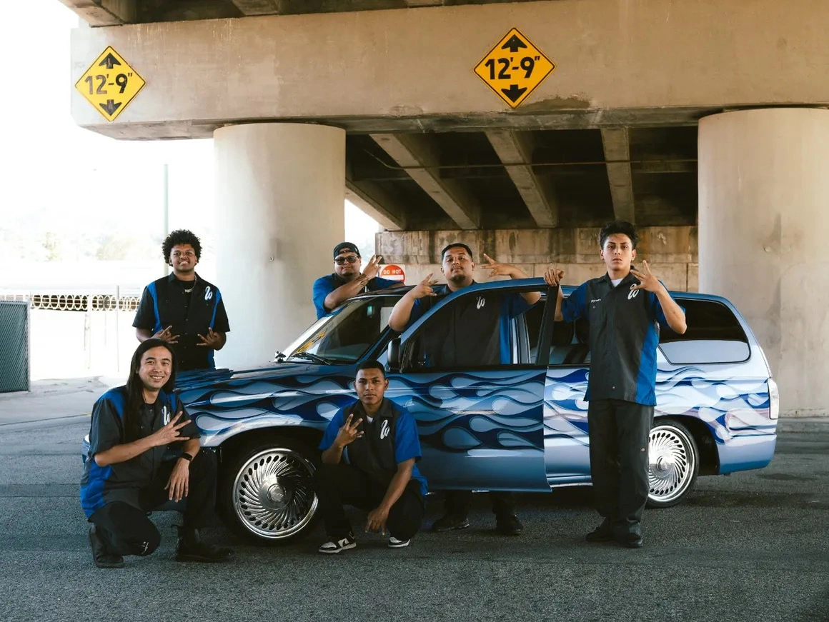 West Coast Customs Academy students posing with a flame-painted lowrider under a freeway overpass