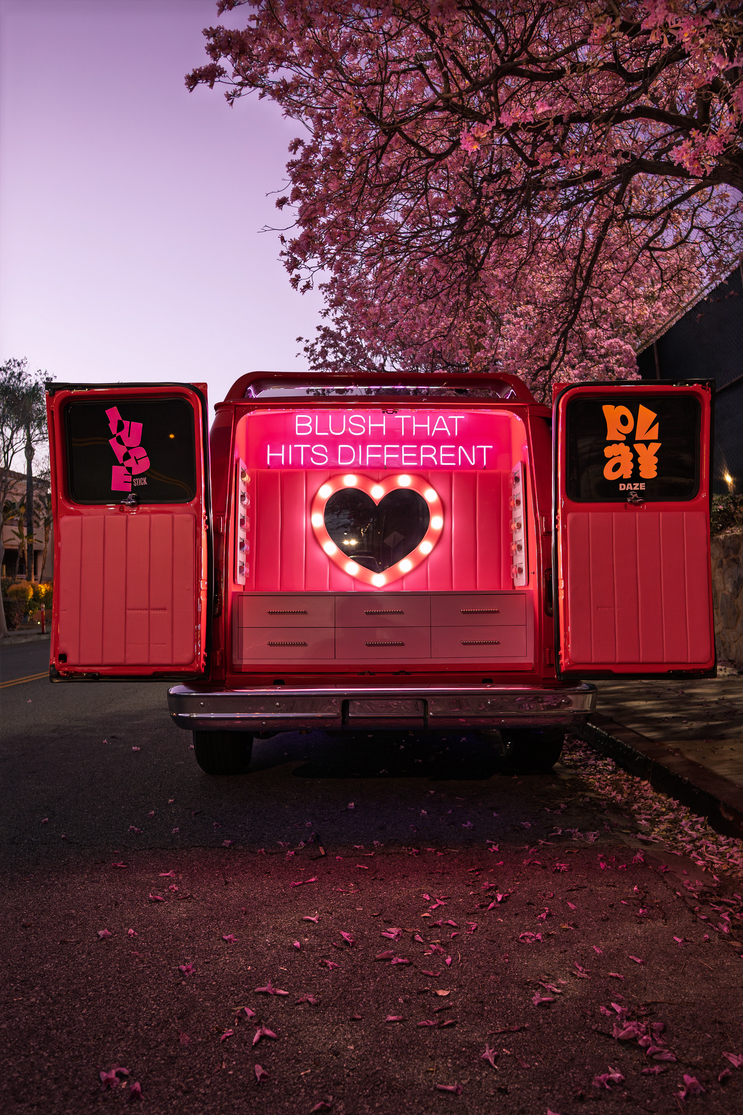 Custom van with its rear doors open to a pink neon “Blush that hits different” vanity under a blossoming tree